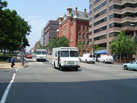 2004 Ford Crown Victoria - Multiple USPS trucks and vans are seen driving on a city street with buildings and trees.