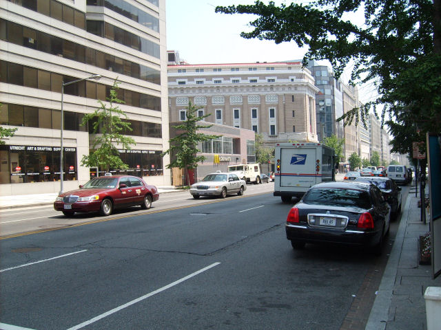 Red sedan and black sedan on a city street with buildings and trees