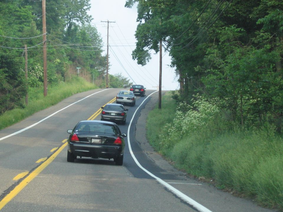 Black sedan from rear, driving on a two-lane road with other cars ahead