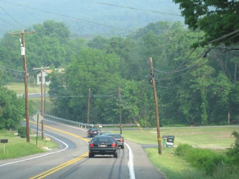 1991 Ford LTD Crown Victoria - A black 1991 Ford Crown Victoria LTD leads a small procession of cars down a winding rural highway, surrounded by lush green trees.