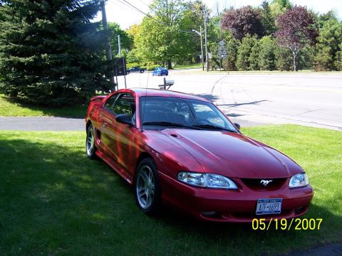 1996 Ford Probe - A red coupe with a horse emblem on the grille is parked on a grassy area next to a road.