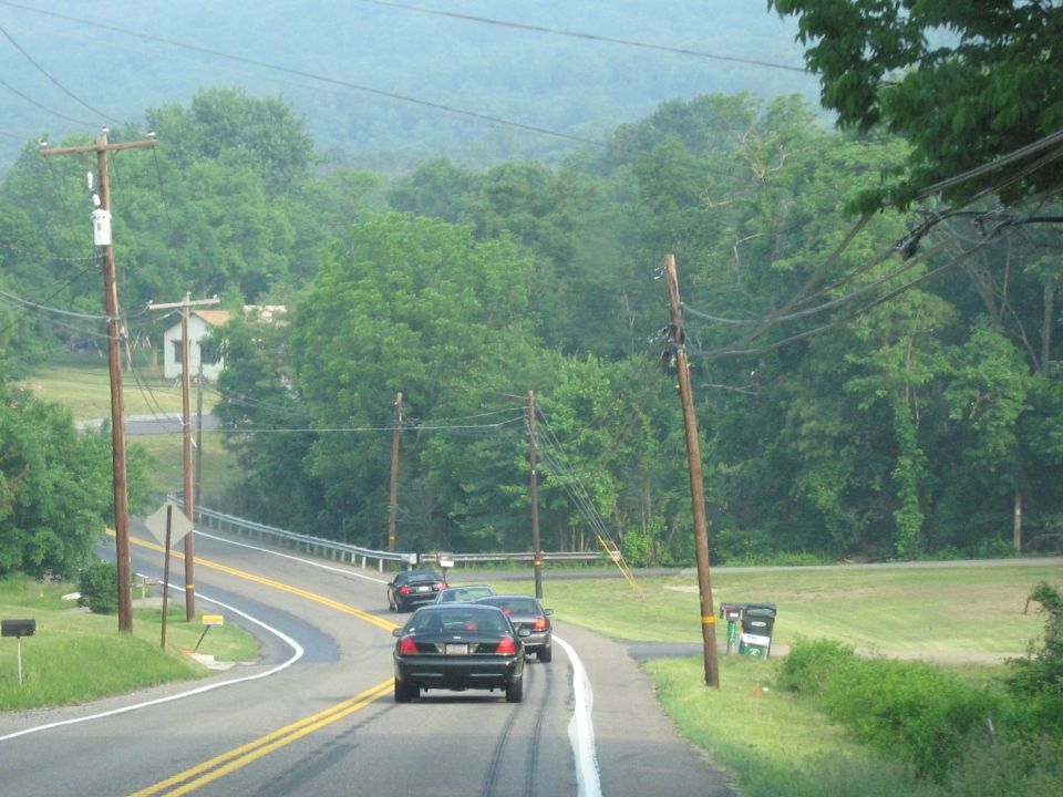 Several cars driving on a two-lane road with telephone poles and trees. One car is in the foreground