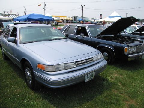 1991 Ford LTD Crown Victoria - A light blue 1991 Ford Crown Victoria is parked next to a dark-colored vehicle with its hood open at an outdoor event.