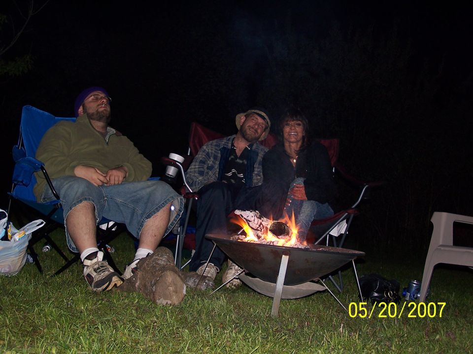 Three people sit in chairs around a campfire at night