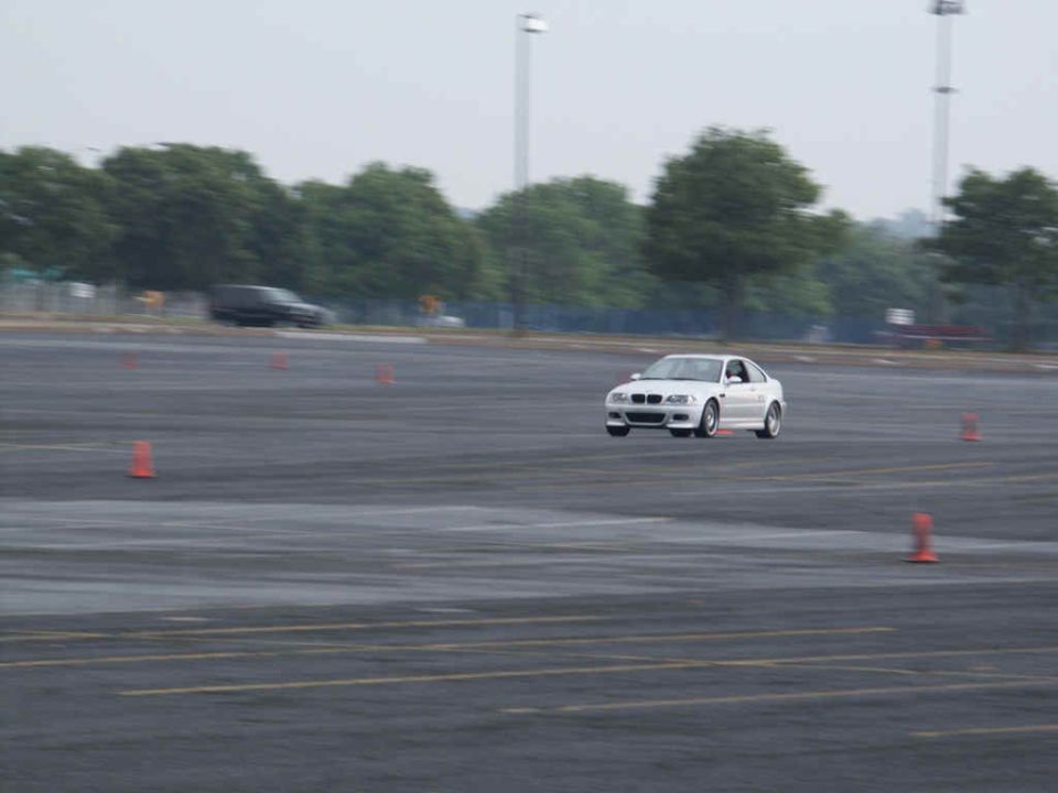 White sedan on a wet asphalt lot with orange cones, blurry background trees and poles. A dark SUV is visible in the distance