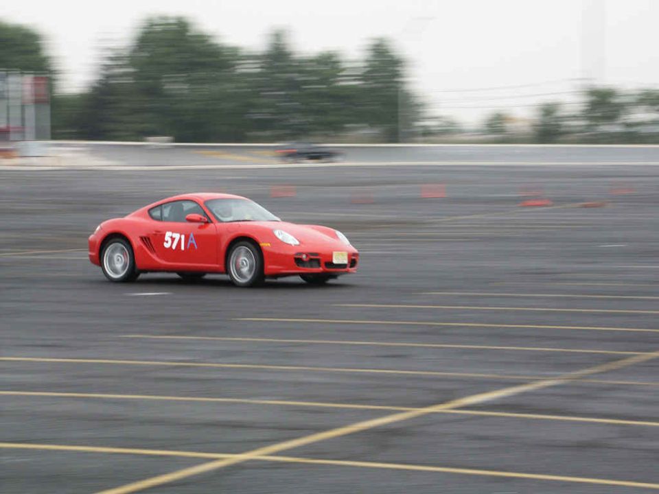 Red Porsche Cayman with number 571A on the door driving on a paved track with orange cones