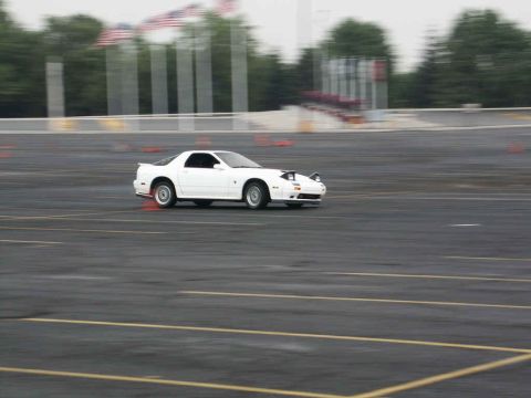 2001 Mercury Grand Marquis - A white sports car is captured in motion on a paved track with orange cones and American flags in the background.