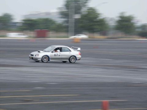 2001 Mercury Grand Marquis - A silver Subaru WRX with a racing number is captured in motion on a paved track, with trees and buildings blurred in the background.