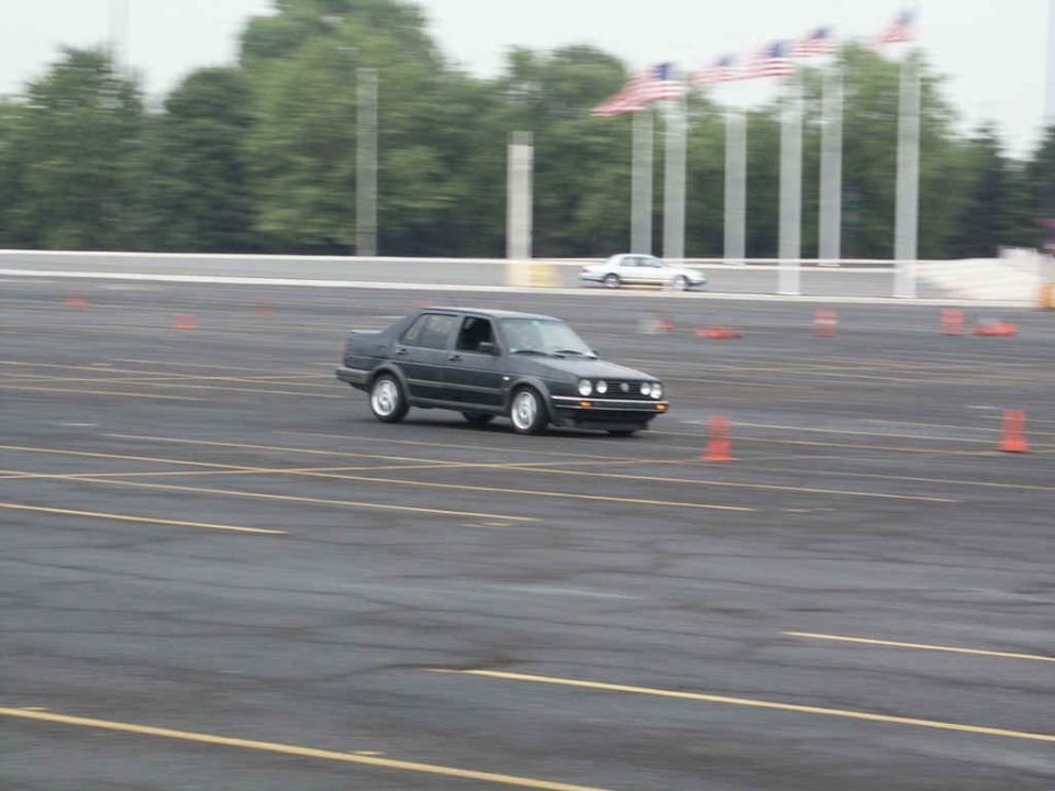 Dark sedan driving on a paved course with orange cones. Another white car is in the background