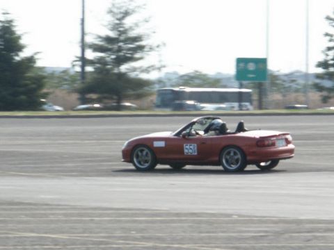 2001 Mercury Grand Marquis - A red convertible sports car is seen on a paved surface, possibly a track, with a bus in the background.