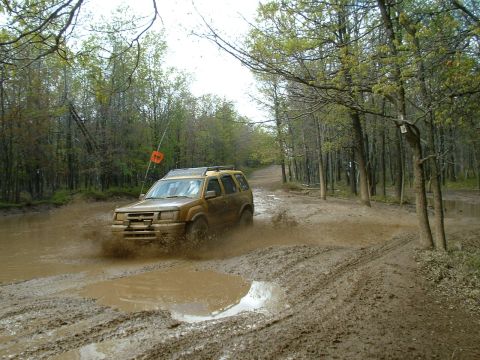1991 Ford Bronco - A gold SUV navigates a challenging muddy trail, kicking up water as it goes.