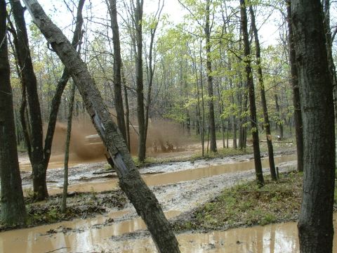 1991 Ford Bronco - An off-road vehicle kicks up a large spray of mud while navigating a waterlogged path through a dense forest.
