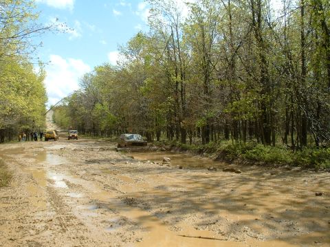 1991 Ford Bronco - Multiple vehicles navigate a challenging muddy road, with trees lining both sides under a partly cloudy sky.