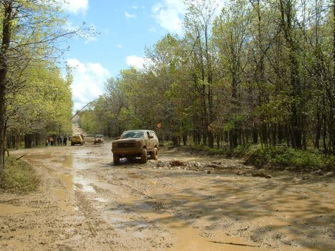 1991 Ford Bronco - Several off-road vehicles navigate a waterlogged dirt path surrounded by lush green trees under a partly cloudy sky.