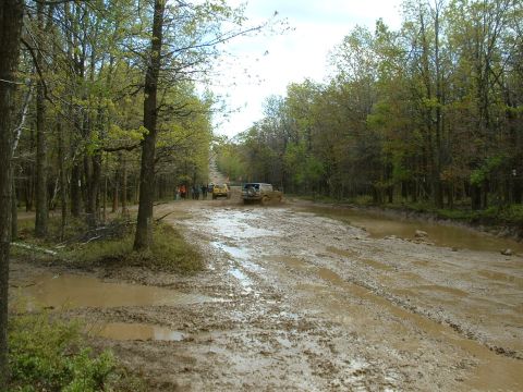 1991 Ford Bronco - Several off-road vehicles traverse a deeply rutted and muddy path through a wooded area.