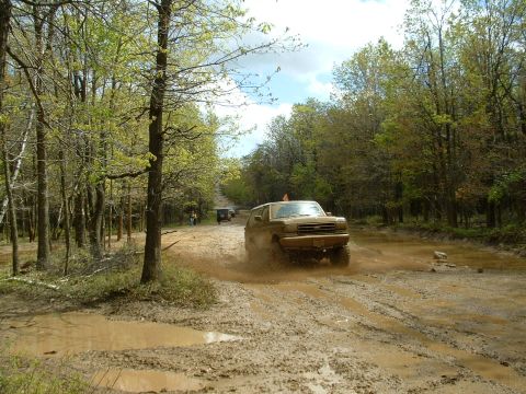 1991 Ford Bronco - A Ford Bronco kicks up mud as it navigates a wet, wooded trail with another vehicle in the distance.