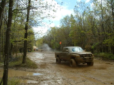 1991 Ford Bronco - A pickup truck navigates a muddy trail surrounded by trees, with other vehicles visible in the distance.