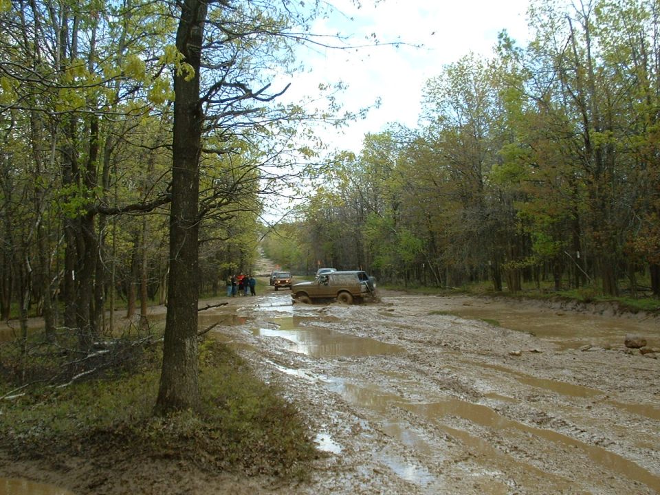 Several SUVs driving through a muddy, wooded trail
