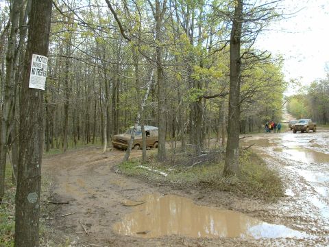 1991 Ford Bronco - A 1991 Ford Bronco navigates a challenging muddy path through a wooded area, with other vehicles visible in the distance.