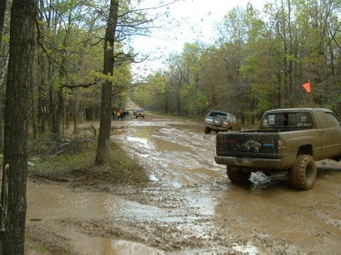 1991 Ford Bronco - Several off-road vehicles navigate a muddy track through a wooded area.
