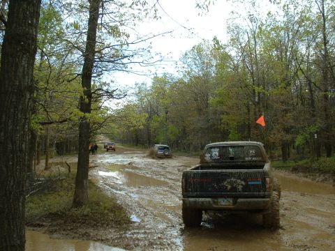 1991 Ford Bronco - Multiple off-road vehicles traverse a muddy trail through a wooded area.
