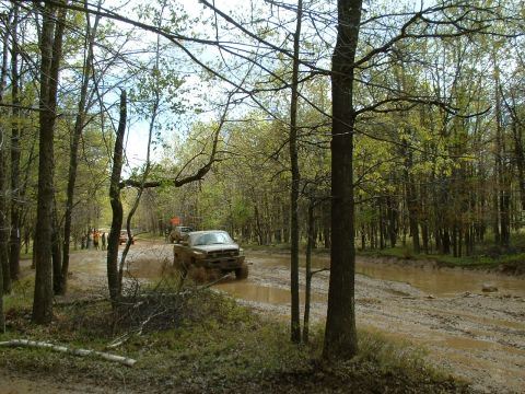 1991 Ford Bronco - Off-road vehicles navigate a challenging muddy trail surrounded by trees.