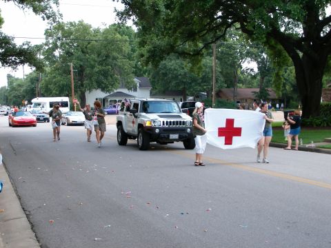 2000 Ford Crown Victoria - A white Hummer H3 leads a parade, followed by people carrying a large Red Cross flag.