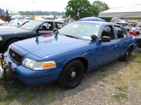 1991 Ford LTD Crown Victoria - A blue police car is parked on a gravel lot with other vehicles and tents in the background.