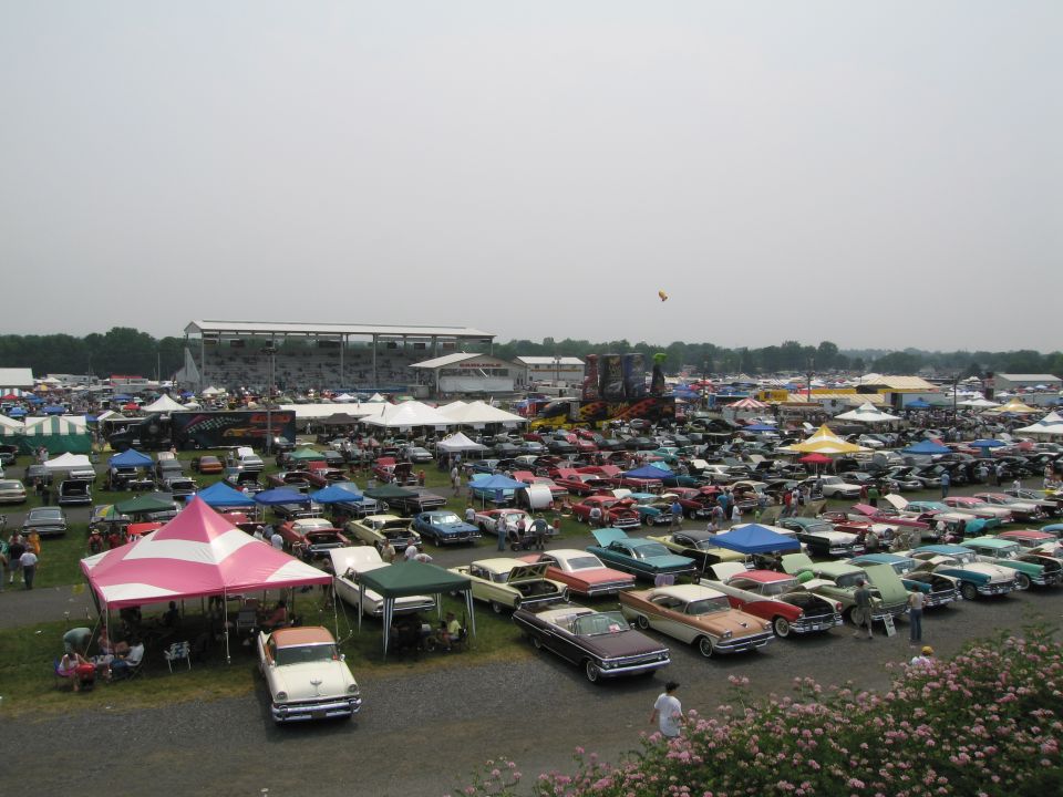 Car show scene with rows of vintage cars parked under tents and open sky