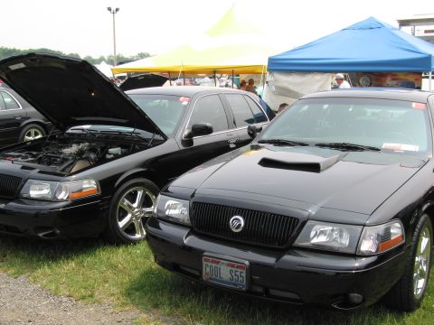 1991 Ford LTD Crown Victoria - Two black sedans are parked on grass at an outdoor event, one with its hood open.