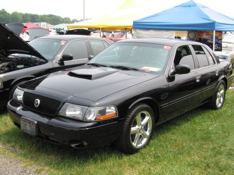 1991 Ford LTD Crown Victoria - A black sedan with a hood scoop and custom wheels is parked on a grassy field next to another vehicle with its hood open.