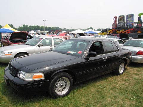 1991 Ford LTD Crown Victoria - A black sedan is parked on a grassy field at an outdoor event with other vehicles and tents in the background.