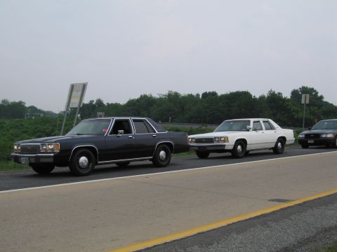 1991 Ford LTD Crown Victoria - Three sedans, including a dark blue and a white Ford Crown Victoria, travel in a line on a highway.