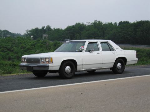 1991 Ford LTD Crown Victoria - This white 1991 Ford Crown Victoria LTD sedan is captured from a three-quarter front perspective on a road with trees in the background.