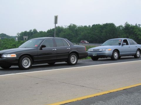 1991 Ford LTD Crown Victoria - Two Ford Crown Victorias are seen on a highway. The black car is on the left, and the light blue car is on the right.