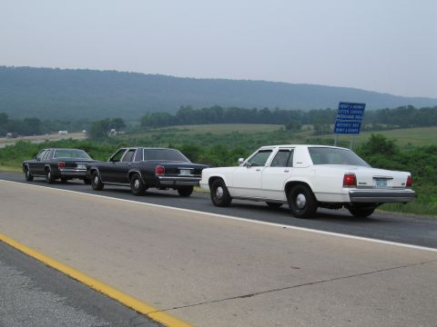1991 Ford LTD Crown Victoria - Three sedans drive in formation on a highway with a scenic, hilly background.