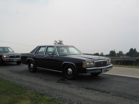 1991 Ford LTD Crown Victoria - A black Ford Crown Victoria sedan is seen from the front-right side, driving on a road with another vehicle behind it.