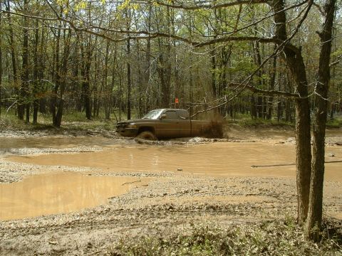 1991 Ford Bronco - A brown pickup truck navigates a muddy obstacle course in a wooded area.