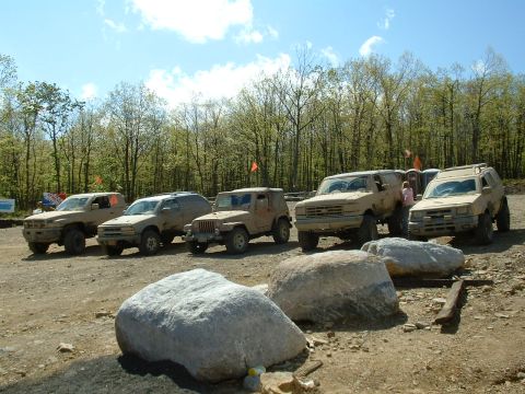 1991 Ford Bronco - A lineup of tan off-road vehicles, including a 1991 Ford Bronco, are parked on a dirt lot with trees in the background.