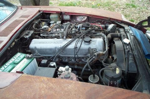 1988 Ford Bronco - The engine bay of a weathered vehicle shows a Nissan OHC engine and a green Interstate battery.