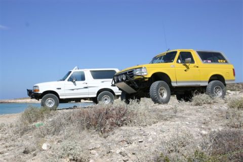 1984 Ford Bronco - Two Ford Broncos, one yellow and one white, are parked on a dry, rocky landscape near the ocean.