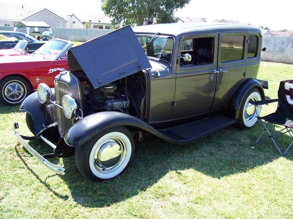 Dark gray vintage sedan with hood open, showing engine. Red car visible to the left