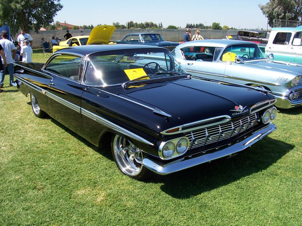 Black 1959 Chevrolet Impala parked on grass, other classic cars nearby
