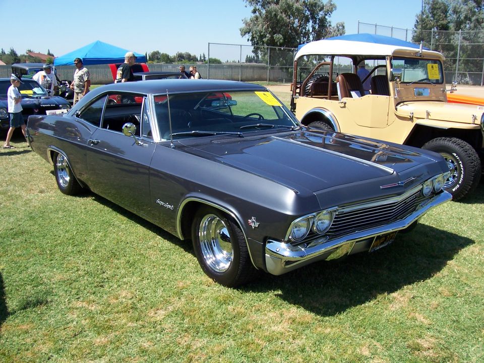 Dark gray 1965 Chevrolet Impala coupe parked on grass, with a tan Jeep-like vehicle behind it