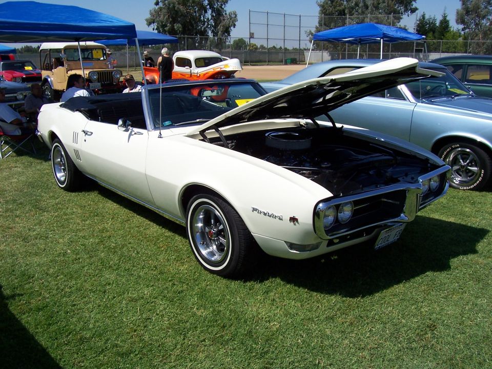 White convertible with hood open, showing engine. Other cars and tents visible in background