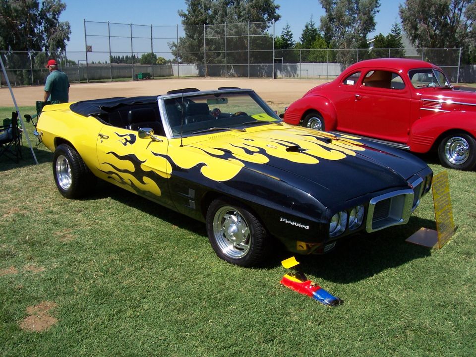 Black convertible with yellow flames, "Firebird" badge visible. Red coupe behind it. Supermotors.net logo in corner