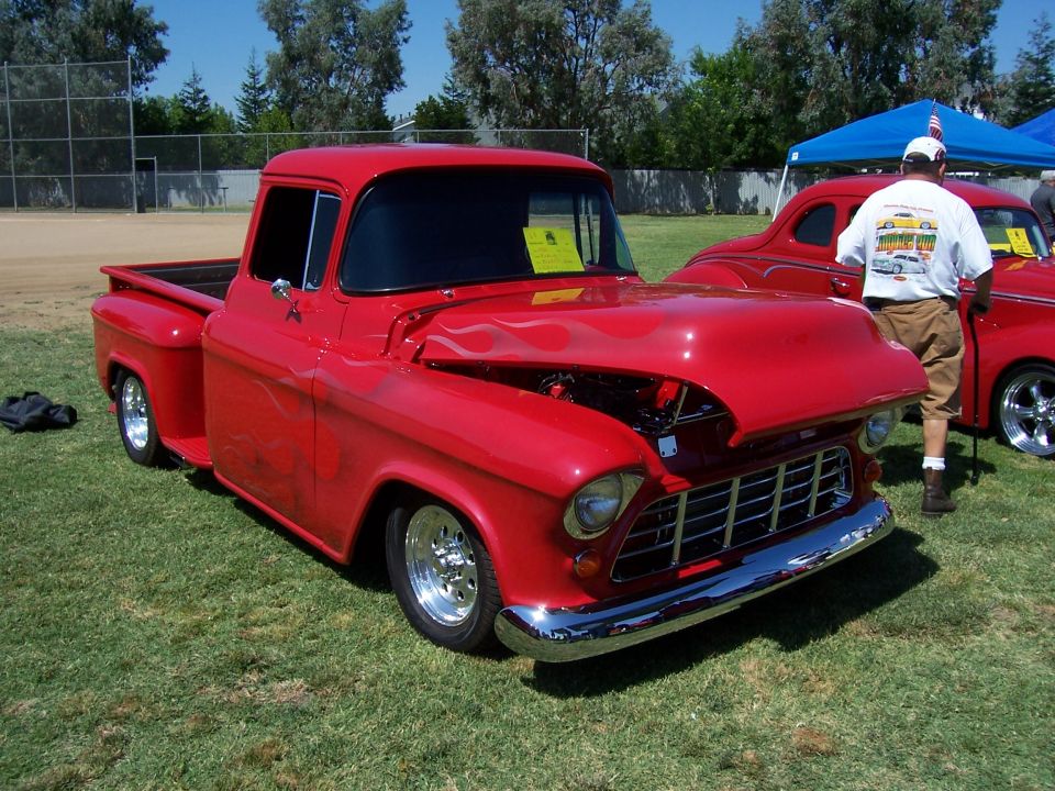 Red pickup truck with flames painted on the hood, parked on grass next to another red car. Hood is open