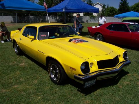 2002 Ford Mustang - A bright yellow 1974 Chevrolet Camaro Rally Sport is displayed outdoors, with a red car visible to its right.
