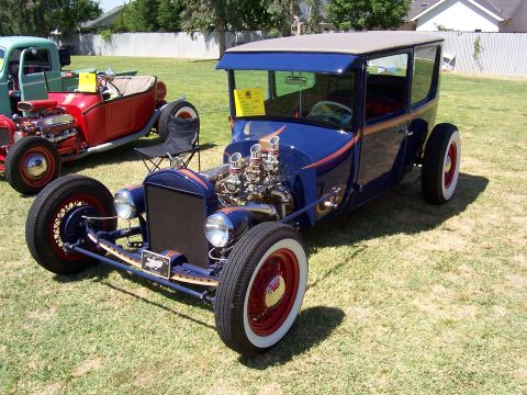 2002 Ford Mustang - A blue hot rod with a prominent exposed engine and red wheels sits on display next to another vehicle.