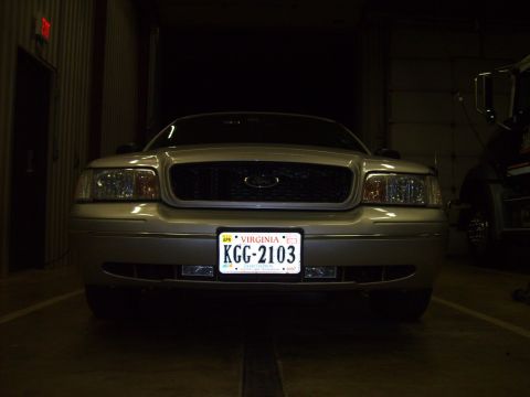2004 Ford Crown Victoria - Front view of a silver/tan Ford Crown Victoria sedan parked inside a dark garage, showing the chrome grille with Ford oval, dual headlights…